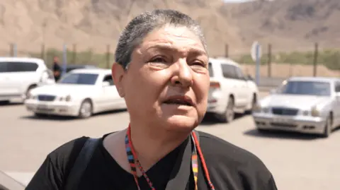 A woman with short cropped hair stands in front of parked cars talking to camera