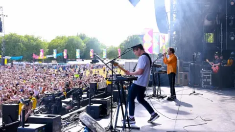 BBC / Sarah Jeynes / Jamie Simonds The Wombats. Three men on a large stage looking out to a large crowd. The stage is open air and two men stand at the front with the third slightly set back.