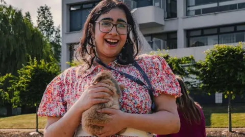 A young woman pictured outside holding a brown rabbit with two hands close to her chest. She is wearing a pink floral dress. She has shoulder-length dark hair and is smiling. 