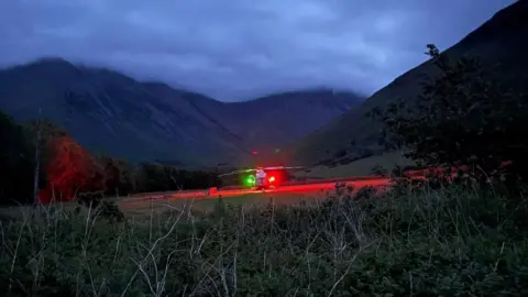 WMRT A helicopter with green and red lights sits on a flat patch of grass in the Wasdale area before setting on a search mission. It is dark and there is thick cloud enveloping the fells. 