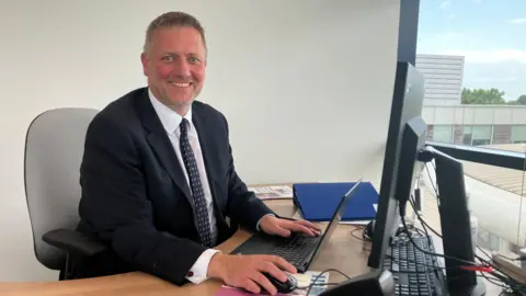 Amy Holmes/BBC A man in a dark suit seated at a desk, using a laptop beside two computer monitors in a bright office with large windows.
