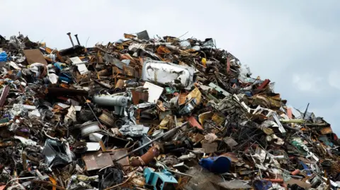 A pile of scrap metal and rubbish lie in a huge pile. There is a clear blue sky above the rubbish pile.