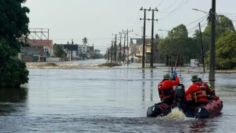 LFB A rescue boat with a team in red life jackets makes its way along a flooded road. Pylons can be seen down in the background.