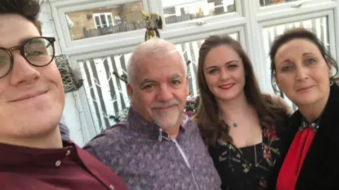 PA A selfie with four people in a row smiling at the camera. On the left is a man with brown hair and glasses wearing a burgundy shirt, then an older man with a grey beard and flowery purple shirt. On the right is a woman with long brown curly hair and red lipstick, and on the end is a woman with dark hair wearing a red dress.