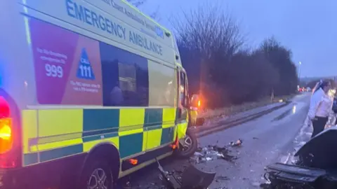 The ambulance involved in the crash is seen from behind. It is on the side of the road and there is crash debris around it and one of the wheels is standing at an angle. The ambulance has its hazard lights on. A man in a white shift is standing near a car on the other side of the road.