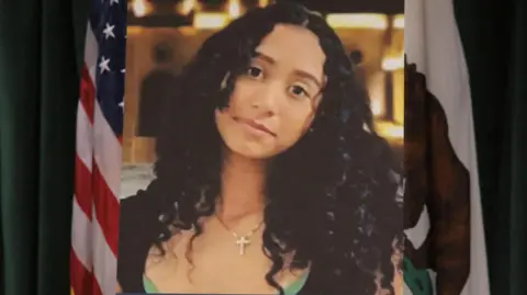 A photo of Celeste Rivas Hernandez is displayed on an easel in front of the US and California flags onstage at a media conference announcing charges against US singer David Anthony Burke, who is known professionally as D4vd.