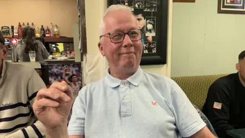 BBC/Lucy Ashton A man with short white hair is wearing glasses and a grey short sleeved top. He is sitting in a social club