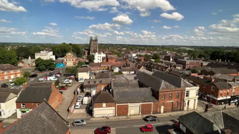 An aerial view of Tamworth, with St Editha's Church the most prominent building, surrounded by rooftops and streets. There are fluffy white clouds in the sky and cars dotted around the streets.