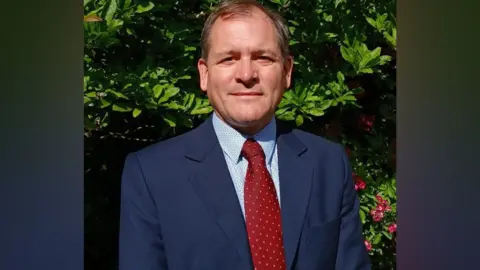 Restore Britain A man looks towards the camera wearing a blue suit, light blue shirt with dots and a red tie in front of a hedgerow