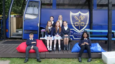 BBC A group of seven pupils are sitting on a bench and some beanbags outside of a blue stationary bus with the school's crest on it.