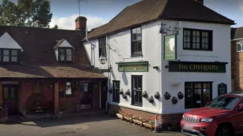 A white building with a green sign which reads "The Chequers" and a red car parked outside