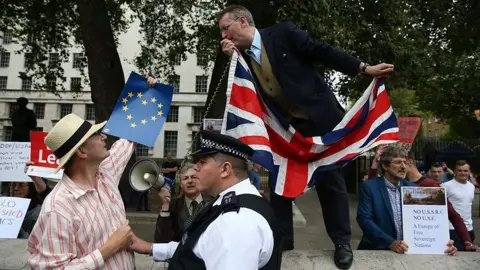 Getty Images A man in a suit stands on a stone ledge holding a Union Jack flag and speaking into a megaphone during an outdoor protest. Below him, a police officer monitors the scene as a demonstrator in a straw hat holds up an EU flag placard with stars shaped like a heart, surrounded by other protesters carrying Eurosceptic political signs in front of a government building.