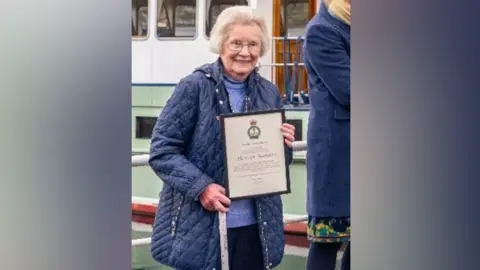 Brenda Couzens is standing holding a certificate and a walking stick. She is wearing a navy quilted jacket and a light blue jumper. She is standing in front of a boat. 
