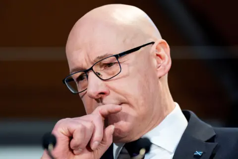 PA Media Head shot of John Swinney, who is bald, holds his right hand up to his mouth and looks at the ground with a serious expression on his face.