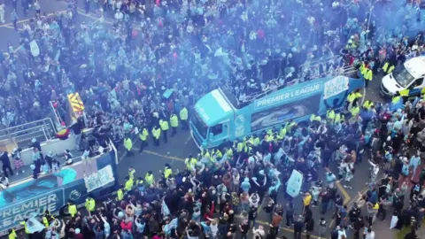 An aerial view of the Coventry City buses. Police officers wearing green hi vis jackets are near the buses and there are large crowds lining the streets. Many are dressed in light blue and a large number have flags. 