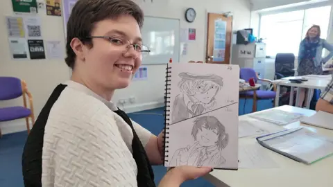 Michael smiles at the camera. He is sitting in a classroom, holding up one of his drawings of two people's faces.