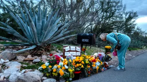 Getty Images A woman in a teal sweatsuit bends over and lays flowers at a memorial for Nancy Guthrie 