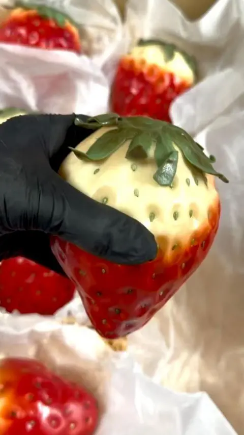 Supplied black-gloved hand holding a giant fake strawberry, which was red at the bottom and yellow at the top, with green leaves sprouting from the head,