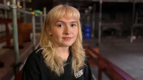 Willow Pearson is a 23 year old woman. She is sitting on a bench with some wooden seating around her. She has long blonde hair and a fringe