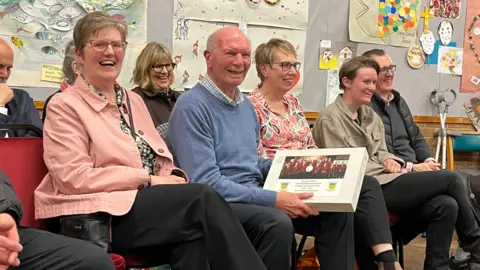 Peter is sat in the middle of the picture smiling and wearing a blue jumper. He is surrounded by people. He is holding a picture of the choir. 