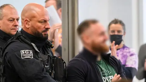 MATTHIAS RIETSCHEL/POOL/AFP Defendant Johann G (blurred and on the right) is brought to the courtroom at the start of a trial against seven left extremists accused for membership in a criminal organization on November 25, 2025 at the Higher Regional Court of Dresden, eastern Germany