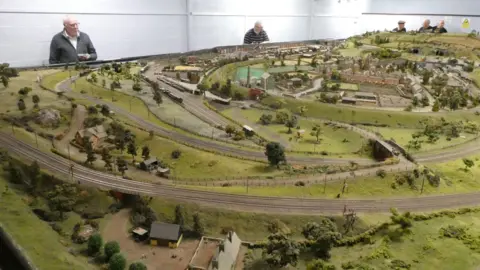 Alan Godber The model railway has green fields, tracks and buildings. There is a white wall in the background. 