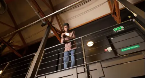 Particle6 Emeka Sesay, a young man, standing and leaning over a balcony. He is wearing blue jeans and a cardigan and white T-shirt.