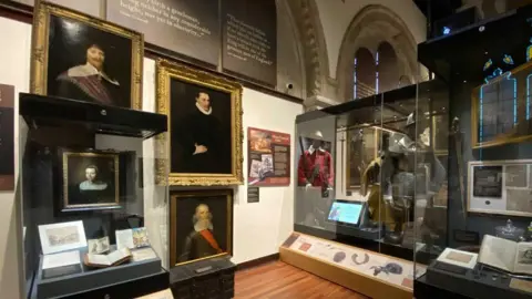 The Cromwell Museum The interior of the Cromwell Museum in Huntingdon. Its white walls have three large gilt-framed portraits of 17th Century men and beneath one is a cabinet showing a small portrait of a 17th Century woman. To the right are glass cabinets showing 17th Century dress and armour, books and documents. 