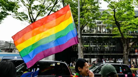 A large rainbow flag - a symbol of LGBTQ rights - flying in the wind, attached to a pole. 