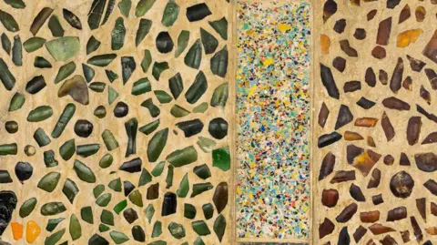 Getty Images Broken glass is used to decorate the walls of Bottle Alley. The glass pieces are collected in shades of green and brown with occasional yellow fragments and there are separate panels decorated in a smaller pattern at intervals.