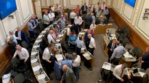 An overhead shot of men and women in a council chamber with crescent-shaped rows of seats