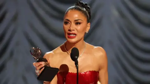 Reuters Nicole Scherzinger accepts Best Performance by an Actress in a Leading Role in a Musical award for Sunset Blvd. at the 78th Annual Tony Awards in New York. she is holding her award and is wearing a red strapless dress.