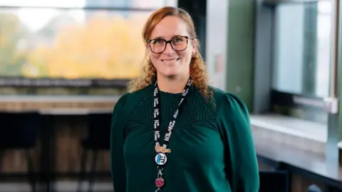 Hull College A woman in a green top smiles at the camera. She is wearing a lanyard and has glasses and long red hair. 