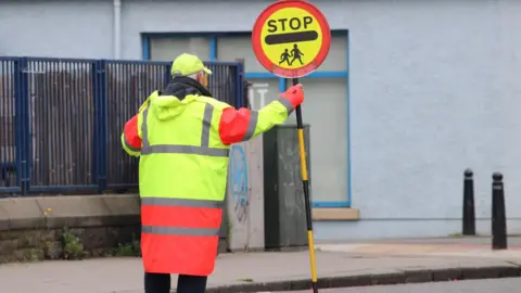 A lollipop man wearing a hi-vis jacket standing in the middle of a road holding a stop sign.