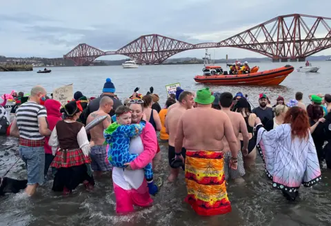 Dozens of people in colourful costumes walk into the Firth of Forth in front of the Forth Rail Bridge. In the foreground is a woman in a pink onesie holding a young boy, wearing a blue onesie.