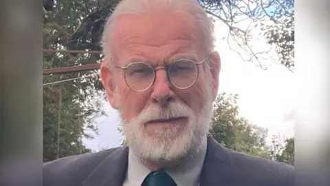 Ulster Architectural Heritage John Anderson, a man with white hair and a matching beard, looks at the camera on a cloudy day. He is wearing a pair of round, metal-framed glasses, a navy jacket, a grey shirt and a dark green tie. There are trees in the background. 