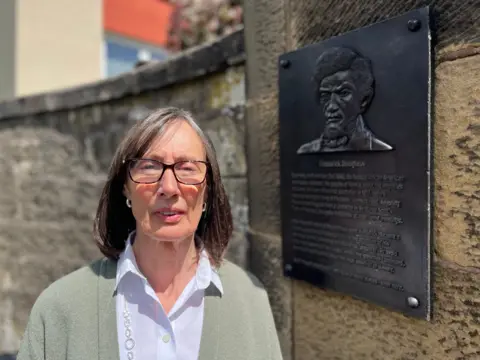 A woman with shoulder-length hair and glasses, wearing a light top and green cardigan, looks at the camera, with a bronze plaque behind her