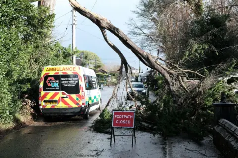 Matt Keeble/PA Wire Fallen tree branch hangs over road. There's a red sign in front reading 'footpath closed' and an ambulance parked to one side of the road
