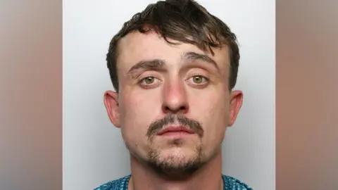 Police mugshot of Kyle Green. He has short brown hair swept over to one side. He has brown moustache and beard.