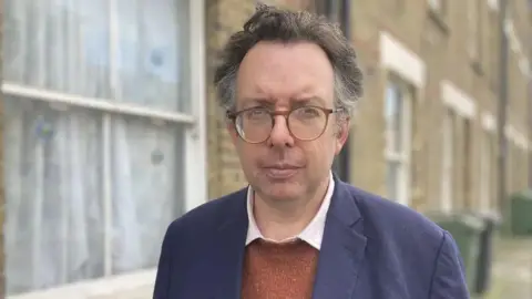 Richard Blakeway is pictured outdoors in front of a row of brick terraced houses. He is wearing glasses, a navy jacket and an orange jumper, looking directly at the camera.