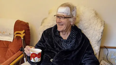 Suzy Weston Brian Mico sitting in a chair wearing his pyjamas and dressing gown, holding a cup of tea. Mr Mico is smiling but has two large white padded dressings on his head after injuries sustained during a fall on the pavements.