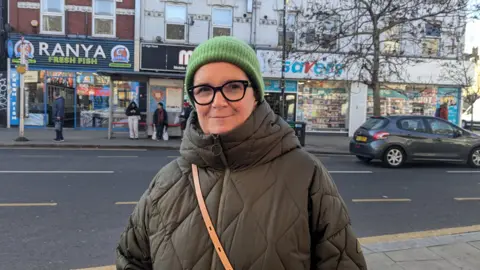 Amanda wearing black framed glasses, a bright green beanie hat, a dark green coat with a thin tan-coloured bag strap across her chest. A road and shops are seen behind her.