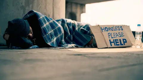 A homeless man in a sleeping bag on a pavement, with a sign asking for help written on a piece of cardboard.