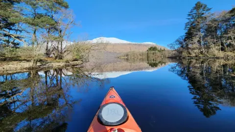 The front of an orange kayak on a very calm Loch Awe. There is a snow-capped mountain in the background reflected in the water which is bright blue, There are trees on either side, also reflected in the water.