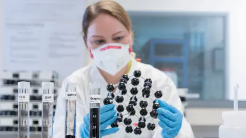 Image shows a female scientist sitting in front of a set of test tubes, and a model of graphene's molecular structure