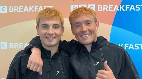 Cian Adams and brother Jordan smile. They are wearing black tracksuit tops and have dyed orange hair. They are standing in front of a BBC Breakfast backdrop sign.