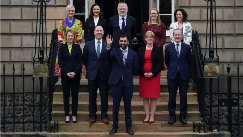 PA Media A group of people stand arranged in rows on the steps of a large stone building, wearing formal clothing, with one individual at the front raising a hand in a greeting gesture.