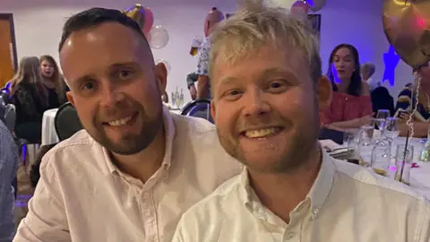 Other Two men in light‑coloured button‑up shirts sit close together at a decorated event with balloons, white‑clothed tables and other guests in the background, both smiling toward the camera.