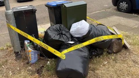 West Northamptonshire Council A large pile of rubbish, with tape round it, next to two wheelie bins and a green metal cabinet. The bin bags and bits of rubbish are on a grass area, with a car to the right.