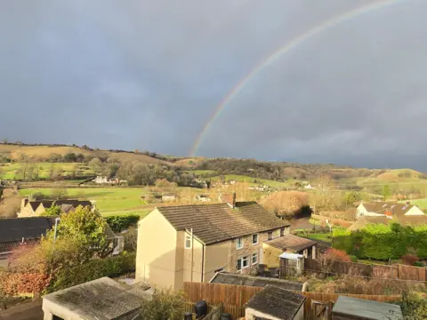 JennyGlos A photo of a row of three houses pictured in front of green hills. The sky is grey and a rainbow appears to be shooting out of the centre of the hills. 